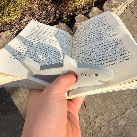 Hand holding a book with a 3D printed grey shark-shaped book holder keeping the pages open outdoors