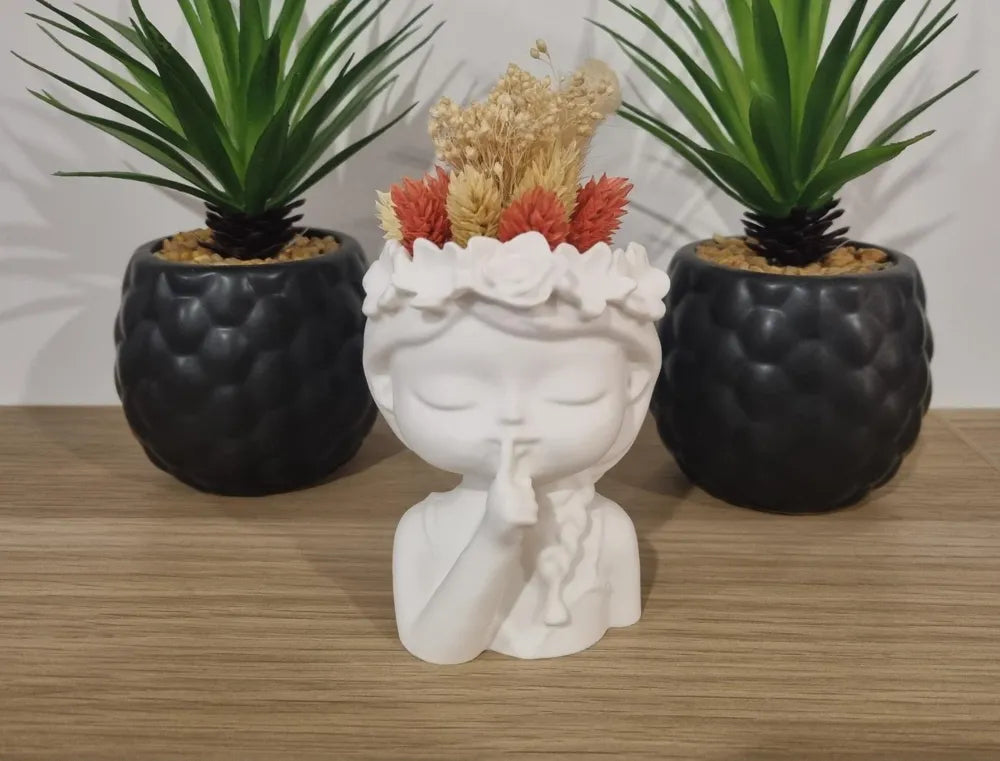 White 3D printed planter shaped like a girl making a shushing gesture with dried flowers inside, flanked by two black textured succulent pots on a wooden surface