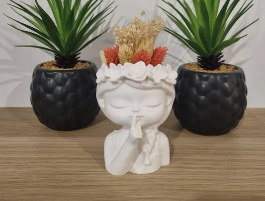 White 3D printed planter shaped like a girl making a shushing gesture with dried flowers inside, flanked by two black textured succulent pots on a wooden surface
